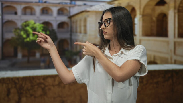 Woman points fingers to the left on an old town street, wearing glasses and white shirt, bare forearms visible; skepticism doubt indecision.