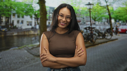 Woman wearing glasses crossing arms on street by canal with parked bicycles, lamp posts and cobblestone pavement  confidence reflection. © Krakenimages.com