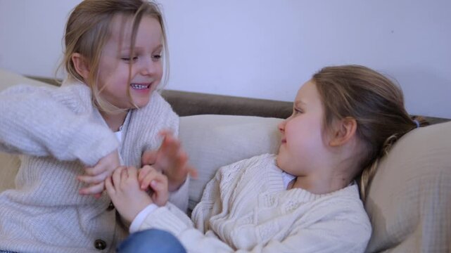 Two cute little sisters having fun together, laughing and tickling each other while sitting on a comfortable couch at home, showcasing a joyful and loving sibling relationship in a cozy atmosphere