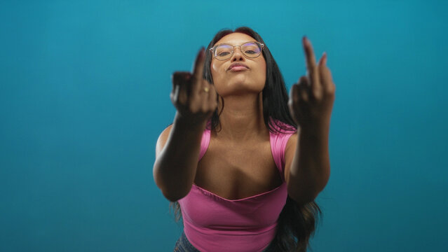 Woman showing both middle fingers with hands extended, wearing pink top and glasses in studio; defiance attitude.