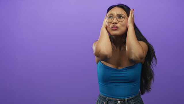 Woman covers ears with hands and winces in a purple studio backdrop wearing blue top and jeans; annoyance sensory overload.