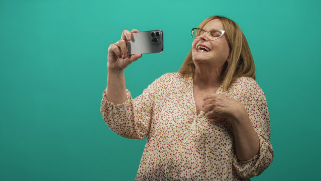 Senior woman wearing glasses and floral blouse holds smartphone with raised arm and hand on chest in studio; joy connection.