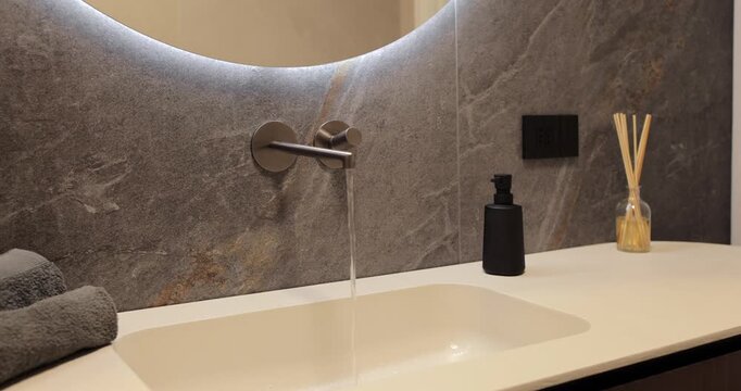 Backlit round mirror above flowing wall faucet and pale integrated basin, static bathroom video presents dark stone surfacing black dispenser reed diffuser and folded towels