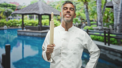 Man chef holds rolling pin upright and hand on hip, smiles with closed eyes beside resort pool in building  contentment. © Krakenimages.com