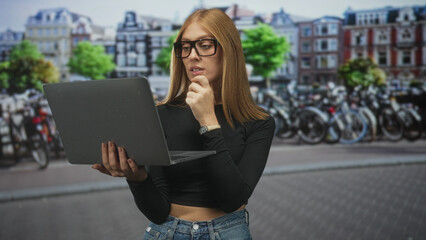 Fototapeta premium Woman holding laptop touching chin with hand on a busy street beside rows of parked bicycles and historic buildings; thoughtful focus.