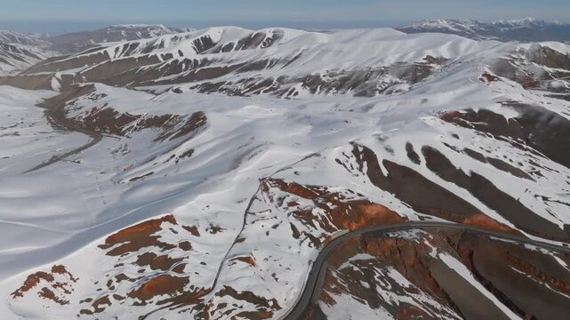 Drone footage of Chyyyrchyk Pass with winding mountain road, snow covered ridgelines and exposed red rock slopes, remote highlands landscape in Kyrgyzstan