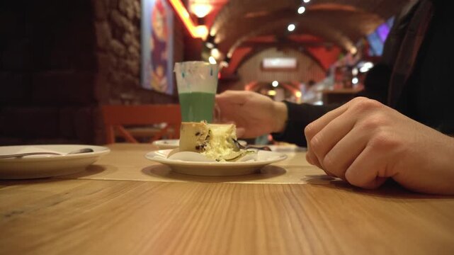 Detailed view of hands of person cutting into a slice of cheesecake, adorned with a lime wedge, placed on a white plate. Blurred interior of cozy restaurant provides warm background. Focus on dessert 