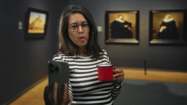 Woman brunette middle aged holding red mug and smartphone makes puckered lips selfie in museum building among framed paintings; playful reflection.