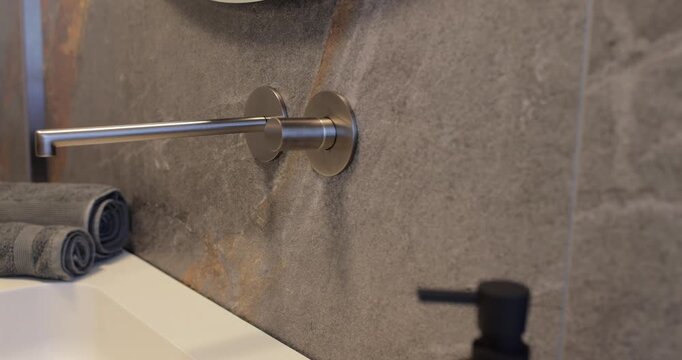 Close view of minimalist wall mounted faucet on dark natural stone, slow camera move tracks across luxury bathroom surface with folded towels soft daylight and blurred black dispenser shaping