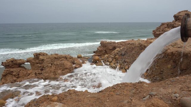 Close-up of a rusty industrial pipe discharging a high-volume stream of wastewater onto a rocky ocean coastline. Perfect B-roll for environmental pollution, coastal erosion, and desalination outfalls.