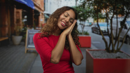 Fototapeta premium Young african american woman with hands together and cheek resting in sleeping gesture on busy city street; restful serenity.