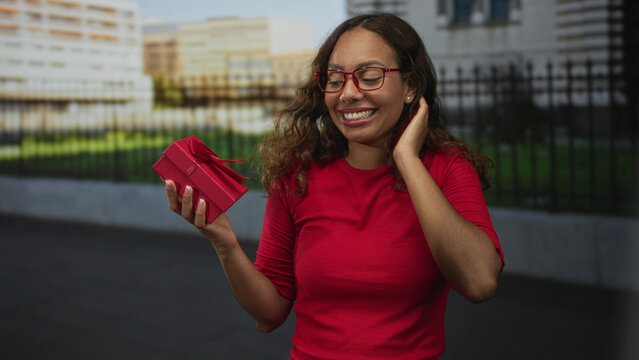 Woman holding red gift box and smiling while touching hair on street near building fence; celebration joy.