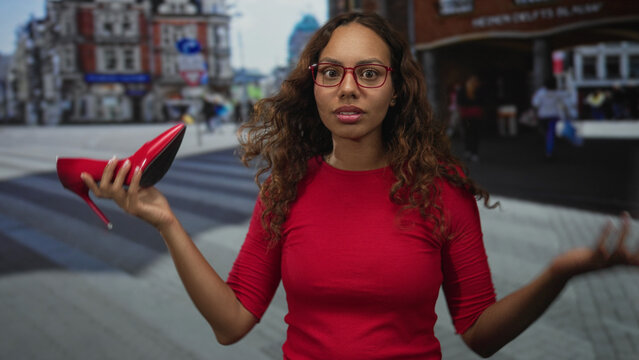 Woman holding red stiletto shoe and shrugging on a busy city street crosswalk; fashion choice confusion.