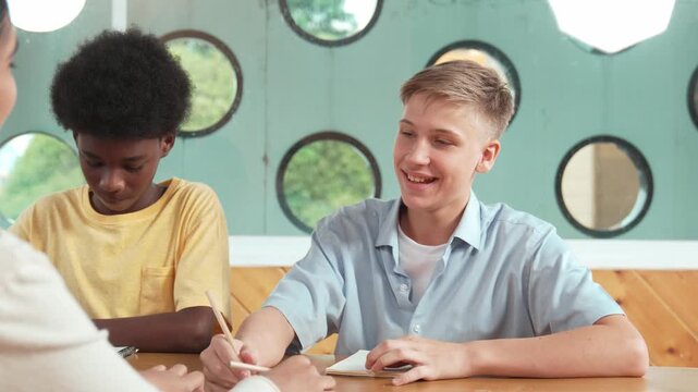 Young smart teenager writing and talking about topic at classroom. Diverse high school student taking a note in other notebook while sitting with multicultural friends. Creative education. Edification