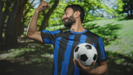 Obraz premium Man bearded soccer player in blue uniform holds soccer ball and flexes bicep while smiling in forest; pride.