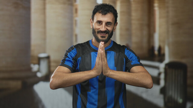 Man in blue and black soccer jersey with hands pressed together praying in building corridor; penitence reflection.