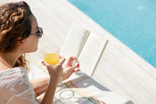 Woman reading book and drinking juice by poolside in summer