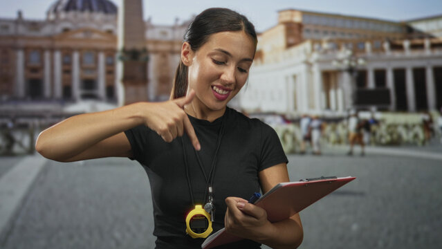 Hispanic woman referee pointing finger down at stopwatch and clipboard in building; confidence coaching.