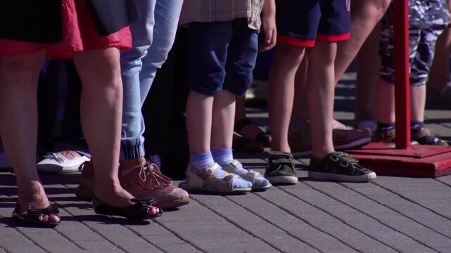 Close-up view of multiple individuals legs and feet, including adults and children, standing in neat rows on a grey paving. Sunlight illuminates variety of shoes and sandals worn by diverse crowd at o
