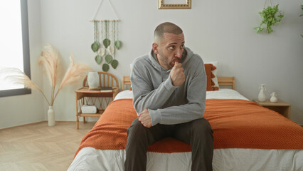 Fototapeta premium Young hispanic man sitting on bed with fist to mouth, wearing gray hoodie, thoughtful pose in studio; quiet contemplation.