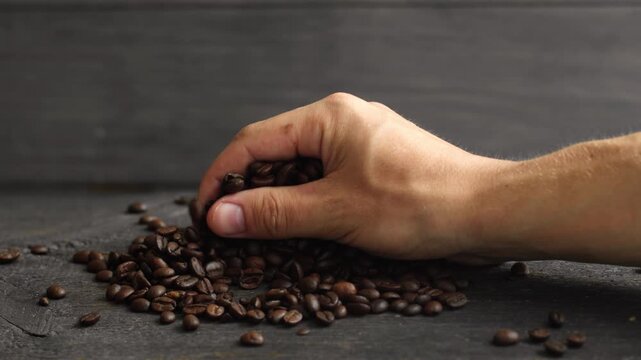 Close-up of a man's hand picking up a handful of aromatic roasted coffee beans and slowly letting them fall back onto a dark table