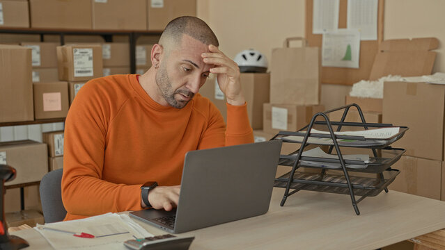 Man writing bills at a desk with laptop and cardboard packages, hand on head gesture in a parcel office building; small business stress.