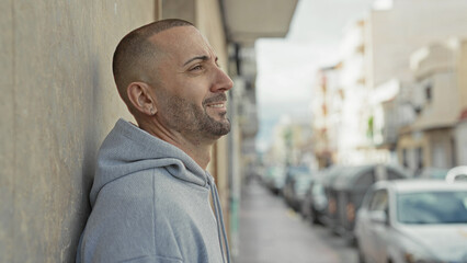 Man leaning against wall, ear visible, smiling with hood up and light stubble, looking up on street  quiet contentment. © Krakenimages.com