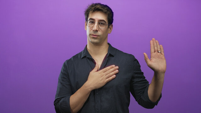 Man wearing glasses and dark shirt, hand placed over chest and other hand raised in oath gesture in studio purple backdrop with wedding ring visible; sincerity.