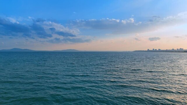 Sunset over the ocean with city skyline. The view shows a wide ocean under a blue sky with some clouds. The city skyline is visible in the distance at sunset.