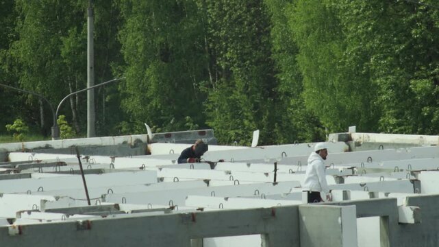 View of construction site with two male workers, one actively welding steel components, creating bright sparks, other worker watching process from distance wearing safety hat, green trees in backgroun