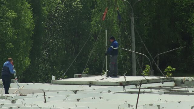 Construction workers building foundation. One worker guides concrete slab by crane. Second worker prepares structure. Outdoor labor, green trees, bright daylight.