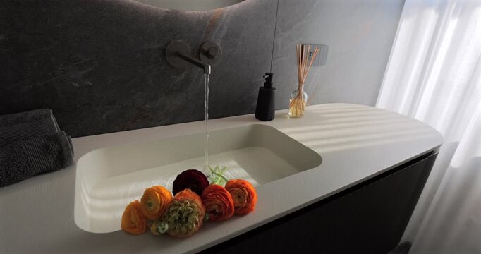 Luxury bathroom sink with running water and fresh flowers, static video of wall mounted faucet filling an integrated basin on dark ribbed vanity beside linen curtains and textured stone wall