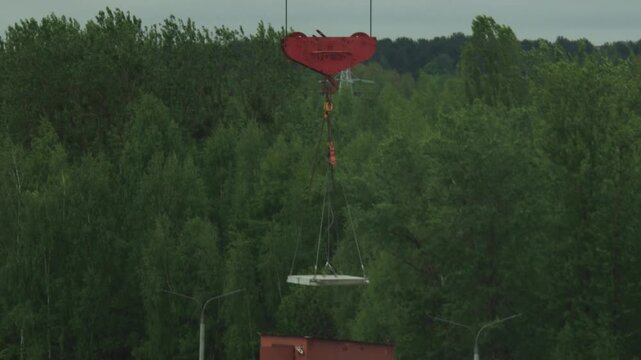 Heavy duty construction crane equipment with large red hook slowly lowering precast concrete slab for building assembly, surrounded by dense forest foliage.