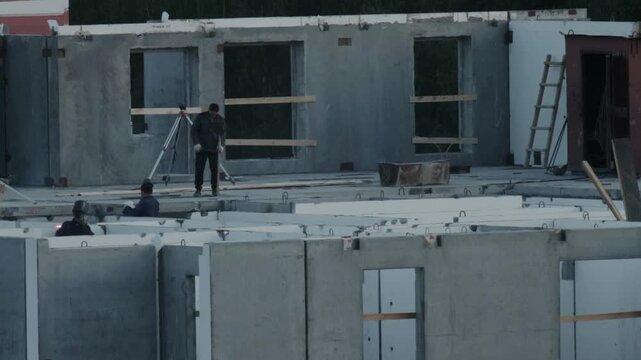 Building construction work by two men. One operates surveying equipment on concrete floor. Another welding concrete panels, sparks visible. Early stage residential structure, raw concrete walls, windo