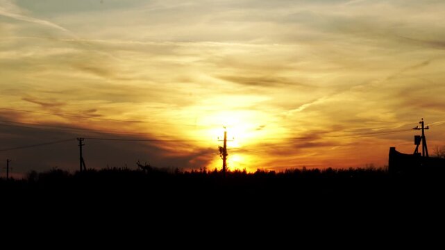 Evening sky displays golden orange and yellow hues of sunset. Silhouettes of power lines, poles, and trees frame horizon. Blurry truck suggests highway traffic, logistics movement.