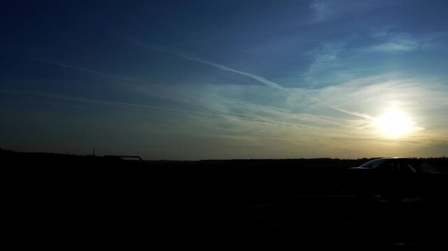 Expansive view of a dark landscape and roadway against a vibrant sunset sky, crisscrossed by multiple airplane contrails. A lone vehicle silhouette adds human element to vast, tranquil scene.