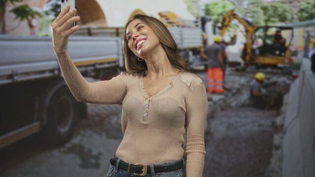 Woman taking selfie with smartphone and raised hand on street beside construction truck and excavator; urban joy.