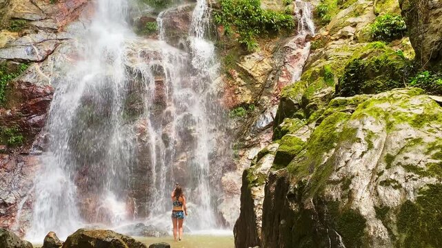A lifestyle shot of a woman in a swimsuit walking through water at the foot of Marinka Waterfalls in Minca. She is using a camera to film the tropical cascades and lush jungle surroundings.