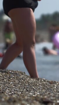 Close-up view of human legs and feet stepping on pebble beach water edge. Blurred background shows people swimming and relaxing, indicating busy summer day vacation.. Vertical shot