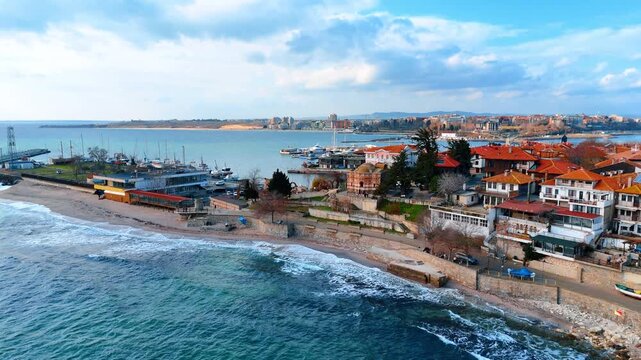 Coastal town with traditional architecture and sandy beach harbor. View of old houses with red roofs along the sea shore with waves hitting the beach and boats in the background.
