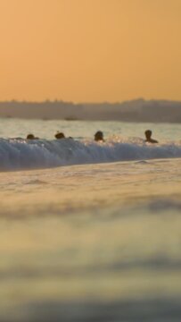Silhouettes of people enjoying ocean water at golden hour, waves breaking on shore with soft focus foreground, warm glow of sunset or sunrise creates tranquil mood on coastal scene. Vertical shot. Sum
