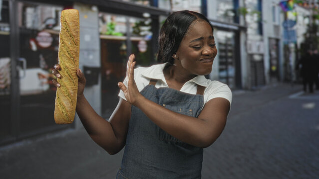 Woman baker in apron holds a baguette in her left hand and recoils with her right hand in a disgusted grimace on a street; disgust hesitation.