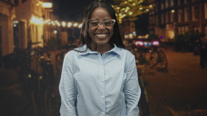 Fototapeta premium Black woman wearing glasses smiles showing teeth on a lit street at night with bicycles and warm string lights; joy confidence warmth.