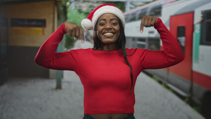 Naklejka premium Woman wearing santa hat points both fingers down at red sweater in a station building, smiling and facing the camera; holiday joy.
