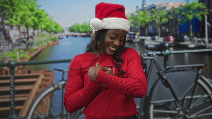 Naklejka premium Woman in santa hat smiles and clasps hands by a canal bridge with bicycle on a street; holiday joy warmth.
