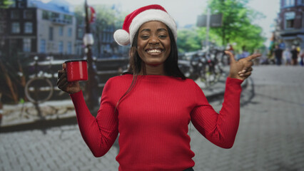 Naklejka premium Woman in santa hat holding a red mug and pointing finger on a cobbled street with bicycles and buildings, wearing red sweater and smiling at camera; holiday joy.