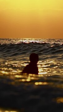 Medium shot of several people swimming and wading in ocean during golden hour. Sun descends on horizon, casting warm orange light across sky and water. Calm, peaceful atmosphere. Vertical shot. Summer