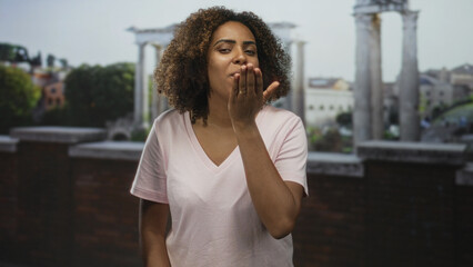 Fototapeta premium Woman blows kiss with cupped hands at ancient roman ruins building outdoors, smiling with hand near mouth; affection travel memory.