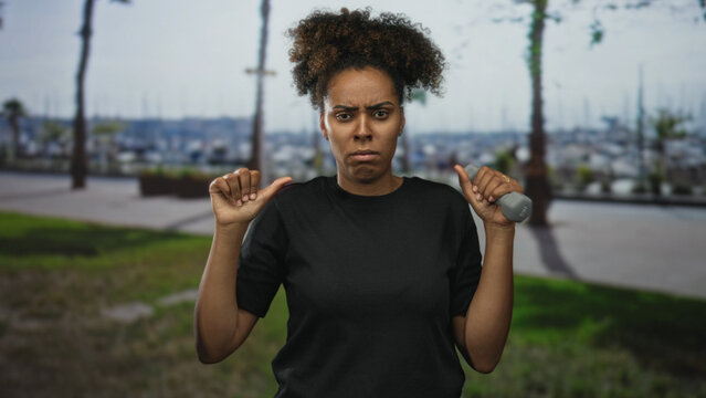 Woman young holding small grey dumbbell with thumbs pointing to chest on street by marina and palm trees near waterfront; defiance.