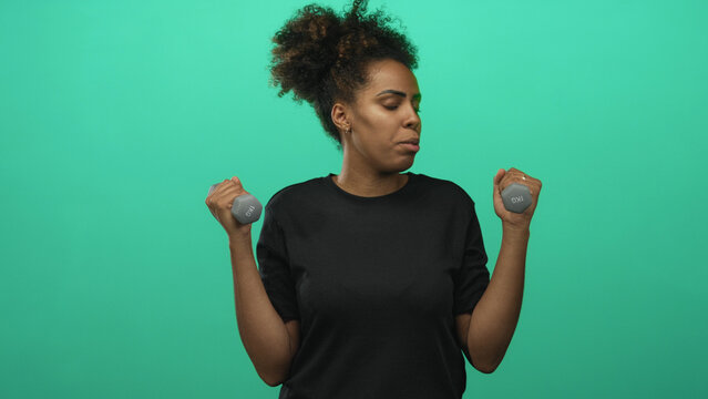 Woman holding small dumbbells and lifting forearms in green studio while wearing black shirt and curly hair pulled up; fitness effort.